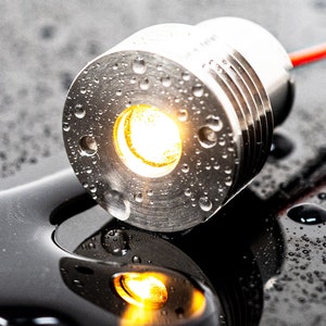 以下が含まれることがあります： 温かい白色の光を放つ、小さく丸い銀色のLEDライト。ライトは水滴で覆われており、黒い表面の上に置かれています。