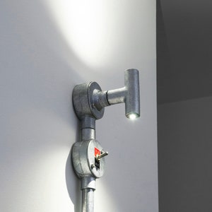Könnte beinhalten: Eine silberne Wandleuchte aus Metall mit einem Schalter. Die Leuchte hat ein Rohrdesign und eine kleine, runde Glühbirne.