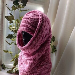 May include: A pink knitted cowl neck scarf with a cable knit pattern. The scarf is folded over and draped around a mannequin's neck.