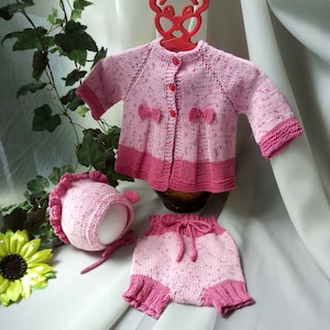 May include: A pink knitted baby outfit set with a cardigan, shorts, and a hat. The cardigan has a button closure and a bow detail. The shorts have a drawstring waist. The hat has a ruffle trim.
