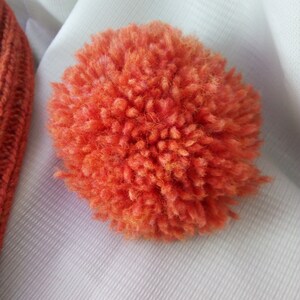 May include: Close-up of a vibrant orange pom-pom made of fluffy yarn. The pom-pom is round and has a dense, textured appearance. Part of an orange knitted hat is visible in the upper left corner. The background is a white fabric.
