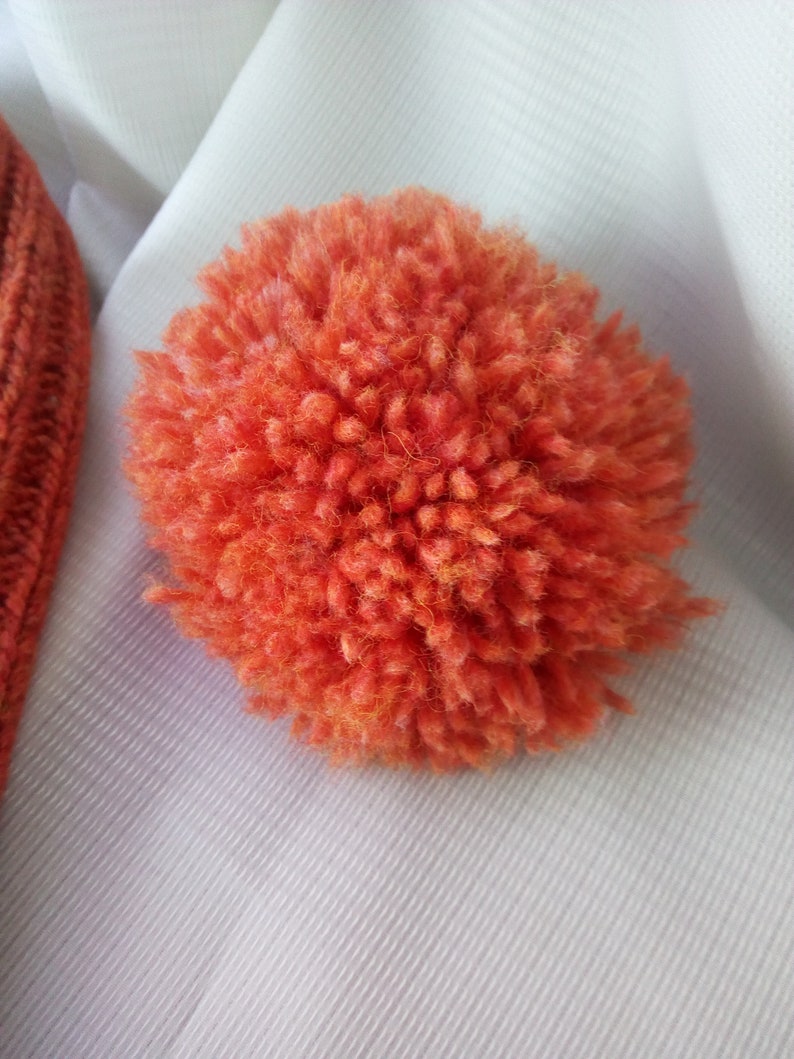 May include: Close-up of a vibrant orange pom-pom made of fluffy yarn. The pom-pom is round and has a dense, textured appearance. Part of an orange knitted hat is visible in the upper left corner. The background is a white fabric.