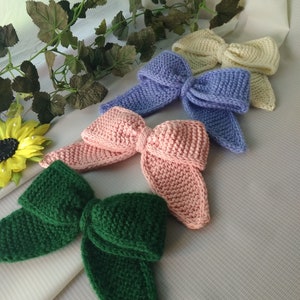 Könnte beinhalten: Vier gestrickte Schleifen in verschiedenen Farben: grün, rosa, lila und creme. Die Schleifen sind in einer Reihe auf einer weißen Oberfläche angeordnet.