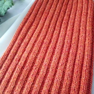 May include: Close-up of an orange ribbed knit hat. The hat is made of a textured yarn and has a vertical rib pattern. The color is a warm, reddish-orange.