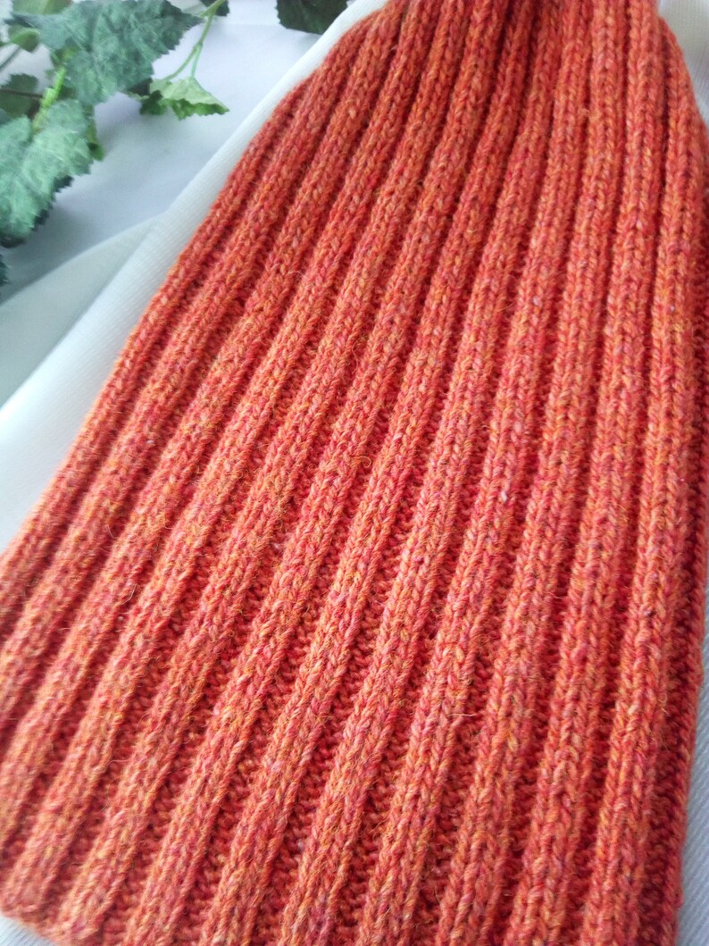 May include: Close-up of an orange ribbed knit hat. The hat is made of a textured yarn and has a vertical rib pattern. The color is a warm, reddish-orange.