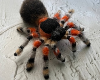 May include: A hand-felted spider with a black and orange striped pattern. The spider has a black and brown body and eight legs.