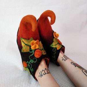May include: A pair of black felt slippers with orange and green pumpkin designs. The slippers have pointed toes and a unique curled shape at the top.
