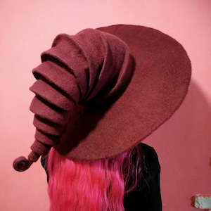 May include: A large, burgundy felt witch hat with a tall, curled brim. The hat is made of a soft, felted material and has a unique, whimsical design.