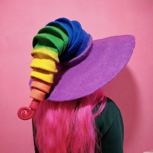 May include: A purple felt witch hat with a rainbow spiral brim. The hat is worn by a person with pink hair.