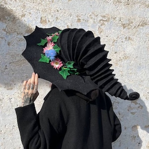 May include: A black felt witch hat with a tall, pleated crown and a wide brim. The brim is decorated with pink and blue felt flowers and green felt leaves.