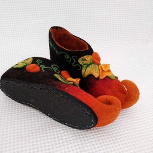 May include: A pair of orange and black felt slippers with a whimsical design featuring green leaves and orange flowers. The slippers have curled up toes and a black leather sole.
