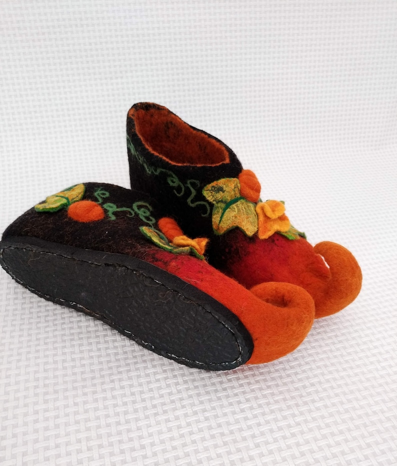 May include: A pair of orange and black felt slippers with a whimsical design featuring green leaves and orange flowers. The slippers have curled up toes and a black leather sole.
