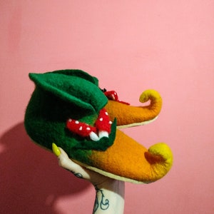 May include: A pair of green and orange felt slippers with pointed toes and a mushroom design. The slippers are held up by a hand with yellow nail polish.