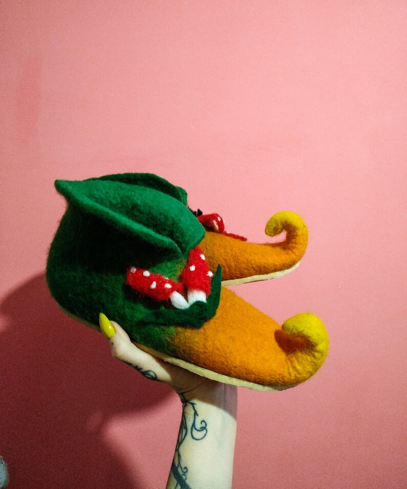 May include: A pair of green and orange felt slippers with pointed toes and a mushroom design. The slippers are held up by a hand with yellow nail polish.