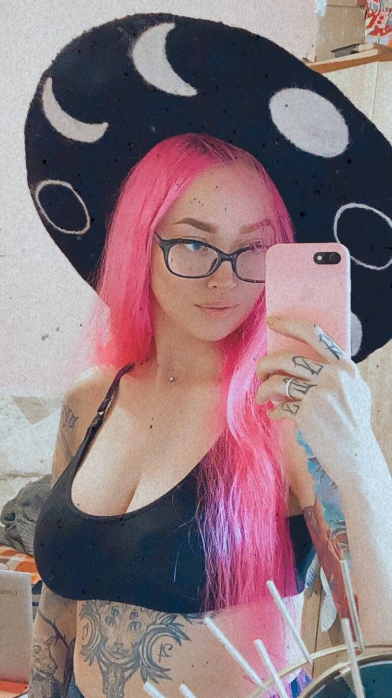 May include: A person with pink hair wearing a black hat with white crescent moons and a black tank top. They are holding a pink phone and have several tattoos on their arms.
