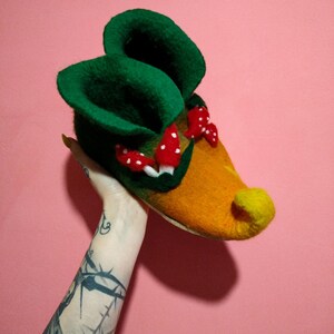 May include: A pair of green and orange felt slippers with red mushroom accents. The slippers are shaped like a gnome's shoes with a pointed toe and a curled up top.