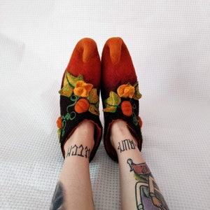 May include: A pair of orange and black felt slippers with a pumpkin design. The slippers have a pointed toe and are decorated with green leaves and orange pumpkins.