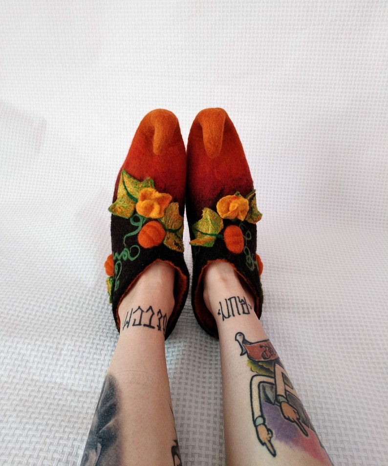 May include: A pair of orange and black felt slippers with a pumpkin design. The slippers have a pointed toe and are decorated with green leaves and orange pumpkins.