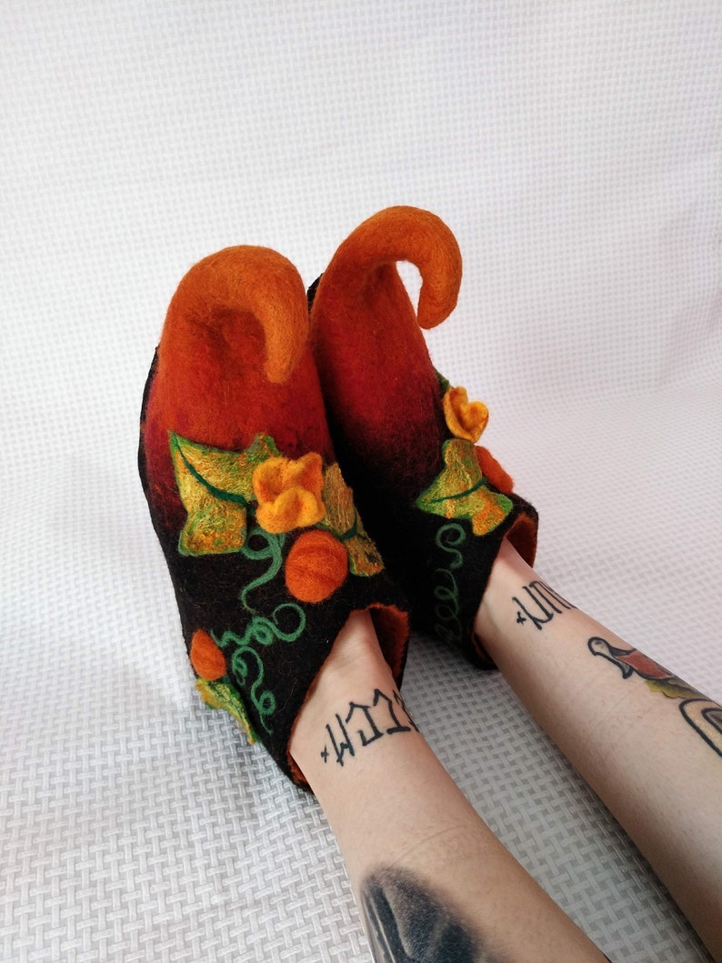 May include: A pair of black felt slippers with orange and green pumpkin designs. The slippers have pointed toes and a unique curled shape at the top.