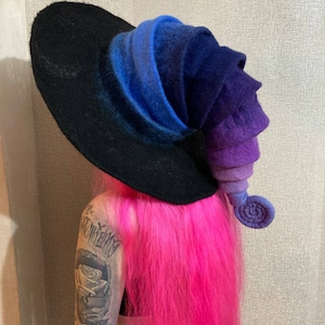 May include: A black witch hat with a blue, purple, and black ombre felt brim and a curled felt top. The hat is worn by a person with pink hair.