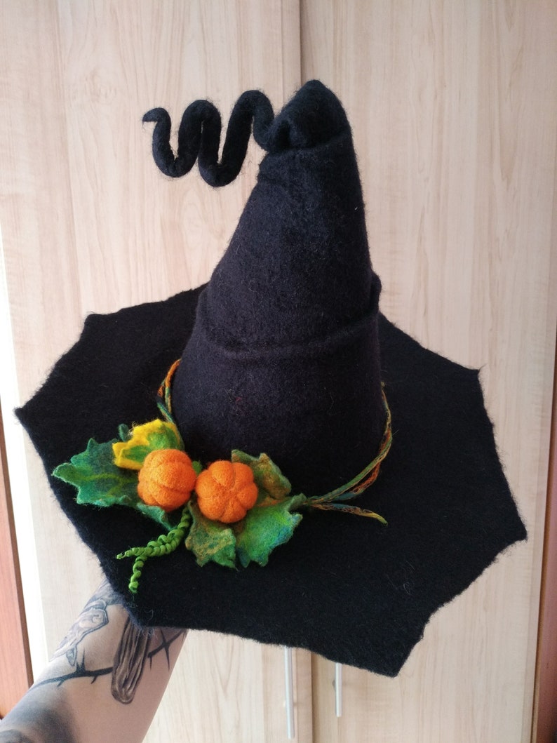 May include: A black felt witch hat with a curled brim and a pointed crown. The hat is decorated with a green felt leaf and two orange felt pumpkins.