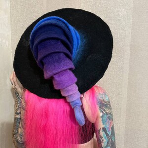 May include: A black witch hat with a wide brim and a tall, pointed crown. The crown is made of felt and is ombre dyed in shades of blue, purple, and pink.