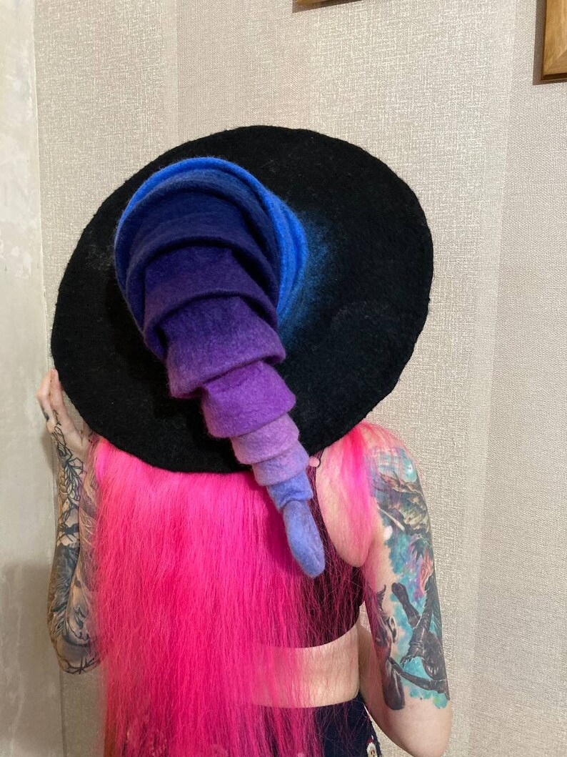 May include: A black witch hat with a wide brim and a tall, pointed crown. The crown is made of felt and is ombre dyed in shades of blue, purple, and pink.