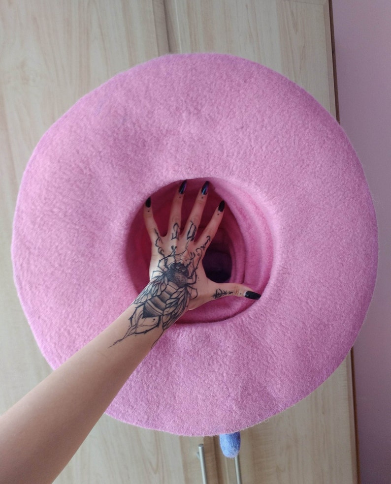May include: A large, pink, felt hat with a wide brim. The hat is being held up by a hand with a black and white bee tattoo.