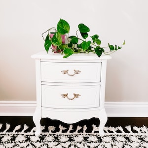 May include: White nightstand with two drawers and ornate gold handles. The nightstand has a curved front and sits on four legs. A green plant is on top of the nightstand.