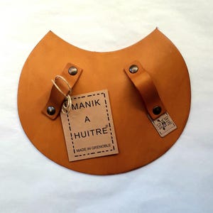 Peut inclure: Un gant à huîtres en cuir tanné avec deux poignées et une étiquette en papier brun qui indique "Manik a Huitre" et "Fabriqué à Grenoble".