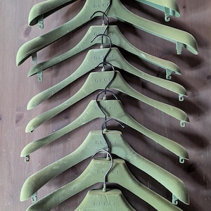 May include: A collection of eight green velvet Gucci hangers with black metal hooks. The brand name "GUCCI" is printed in a matching green color on each hanger. The hangers are arranged vertically against a brown wooden background.