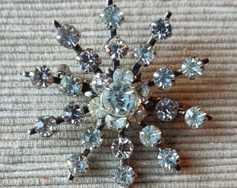 May include: A silver snowflake brooch with clear rhinestones. The brooch has a starburst design with a central cluster of rhinestones.