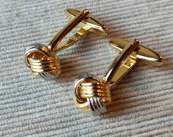 Puede incluir: Dos gemelos de oro con un diseño de nudo. Los nudos están hechos de alambre de oro y plata.