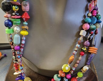 May include: A multi-strand necklace with a variety of colorful beads in different shapes and sizes. The beads include round, oval, and geometric shapes in shades of pink, blue, green, yellow, and orange. The necklace is displayed on a mannequin.