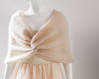 May include: A light beige knitted shawl draped over a mannequin. The shawl is knotted at the front, creating a soft, textured appearance. The mannequin is wearing a light-colored dress with a tulle skirt, suggesting a bridal or formal wear theme.