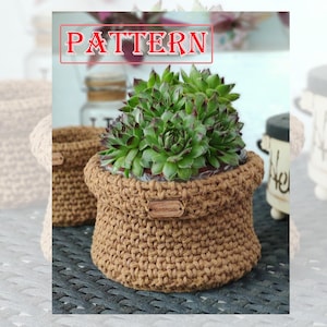 May include: A brown crocheted plant pot with a wooden tag that says "Handmade" and a succulent plant inside. The pot is sitting on a woven wicker surface.