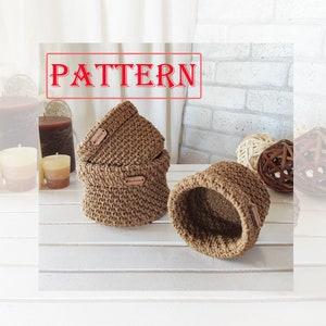 May include: Three brown crocheted baskets with wooden labels. The baskets are stacked on a white surface.
