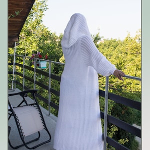 May include: A white knitted cardigan with a hood, long sleeves, and a ribbed texture. The cardigan is worn by a person standing on a balcony overlooking a green landscape.