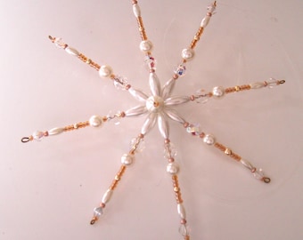 Pode incluir: Um ornamento em forma de estrela composto por contas ovais brancas, pérolas redondas brancas, cristais facetados transparentes e pequenas contas douradas. O design apresenta oito pontas radiantes que se estendem a partir de uma pérola central.