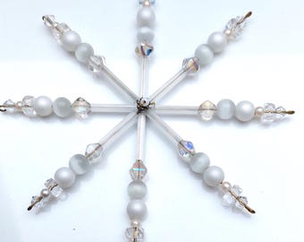 Pode incluir: Um ornamento em forma de floco de neve composto por oito raios metálicos. Cada raio é decorado com uma sequência de contas brancas foscas, contas de cristal facetado transparente e pequenas contas de vidro iridescente, centradas em torno de um eixo metálico.
