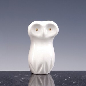 Peut inclure: Figurines de hibou en céramique blanche avec des yeux dorés. Le hibou est debout sur une surface noire.