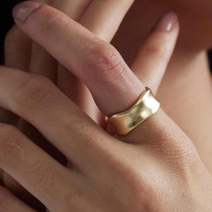 May include: A gold-colored ring with a wavy design. The ring is worn on a finger, and the image is a close-up shot. The ring has a matte finish and a unique, flowing shape, suggesting a modern jewelry style.