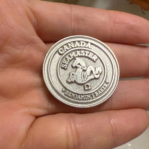 Puede incluir: Una moneda redonda de color plateado con el texto "CANADA SEAMASTER" en el borde superior. La moneda presenta un diseño de caballito de mar en el centro y el nombre "BENJAMIN J BAKER" en la parte inferior. La moneda se sostiene en una mano.