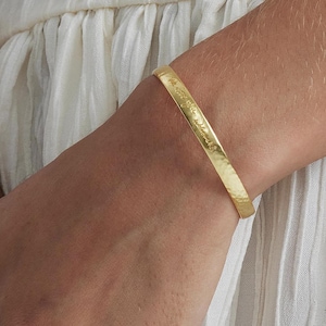 Op de afbeelding: Een goudkleurige manchetarmband met gegraveerde tekst. De armband wordt om de pols van een persoon gedragen en de achtergrond omvat witte stof. De armband heeft een gehamerde textuur.