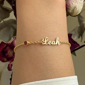 May include: Gold chain bracelet with a small red gemstone and the name "Leah" in cursive.