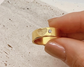 May include: A gold band ring with a fingerprint design in the shape of a heart and a small diamond accent.