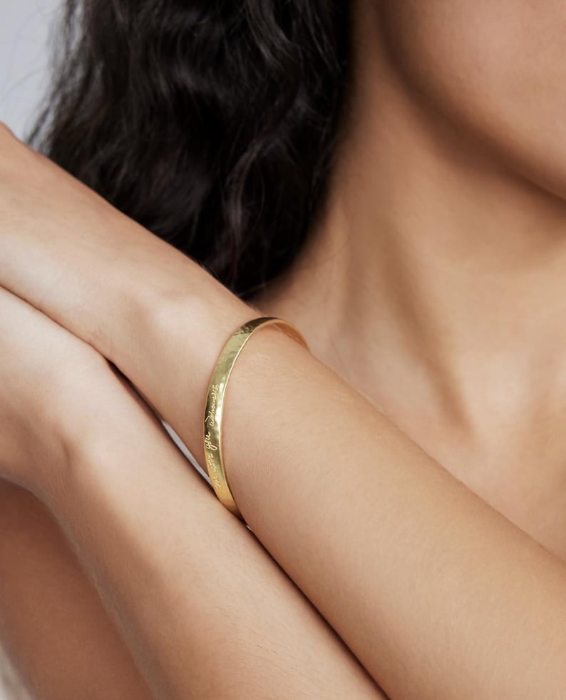 Op de afbeelding: Een goudkleurige armband met gegraveerde tekst. De armband wordt om een arm gedragen. De armband heeft een glad, gepolijst oppervlak en lijkt van metaal te zijn. De tekst is klein en cursief.