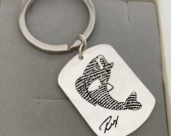 May include: Silver keychain with a black and white engraved image of a fish jumping out of the water. The keychain has a signature that says "Rex".