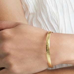 Puede incluir: Un brazalete de color dorado con una textura martillada. El brazalete tiene una inscripción grabada. La joya se lleva en la muñeca, sobre un fondo de tela blanca.