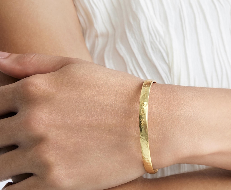 Op de afbeelding: Een goudkleurige manchetarmband met een gehamerde textuur. De armband heeft een gegraveerde inscriptie. De sieraden worden om een pols gedragen, tegen een witte stoffen achtergrond.